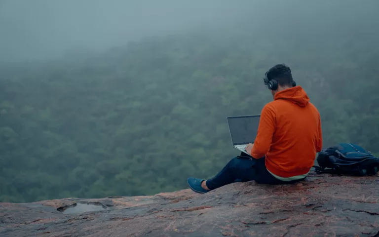 Man in Orange Shirt and Blue Denim Jeans Sitting on Brown Rock Using a Laptop